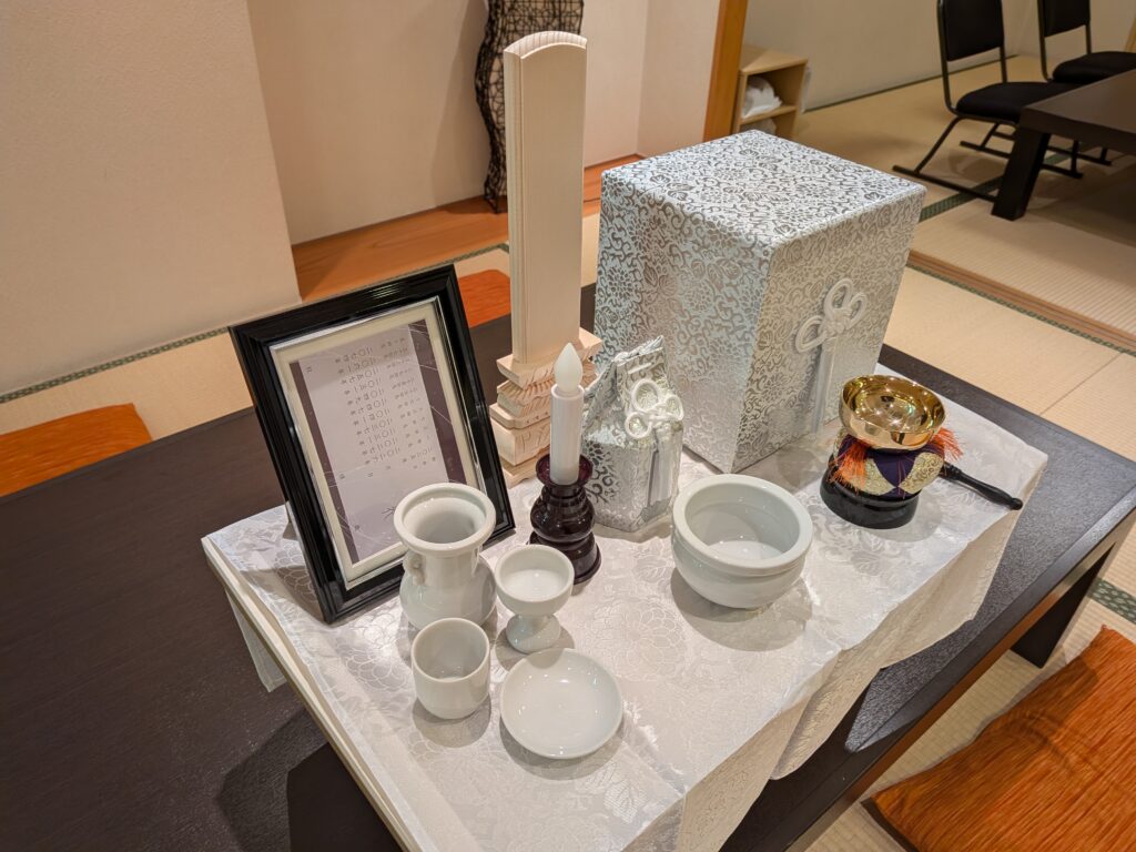 堺市の葬儀で使われる後飾り祭壇に、中型の骨壺と白木位牌、陶器の供物具が整えられた様子が写っている。